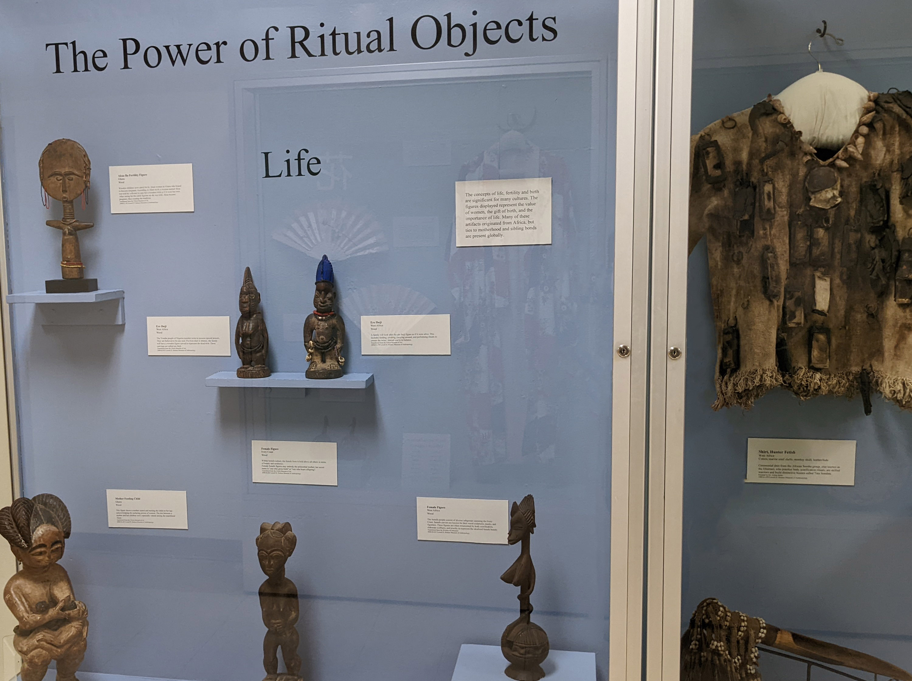 Exhibit with a blue background with small figures and a shirt on display. Title reads "The Power of Ritual Objects: Life"