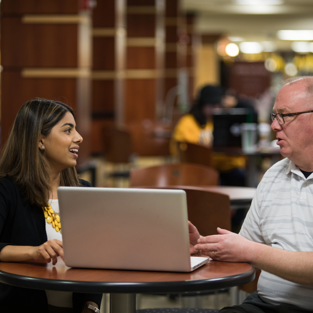 A student and an advisor sit at a small round table with a laptop between them, talking and smiling during a one-on-one advising conversation in a campus common area.