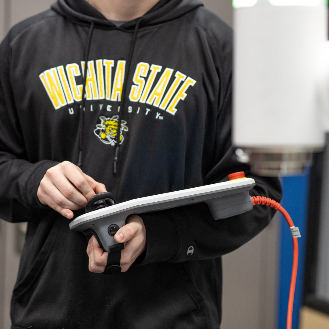 A student in a Wichita State University hoodie operates a handheld control panel connected to lab equipment, hands-on with technical machinery.