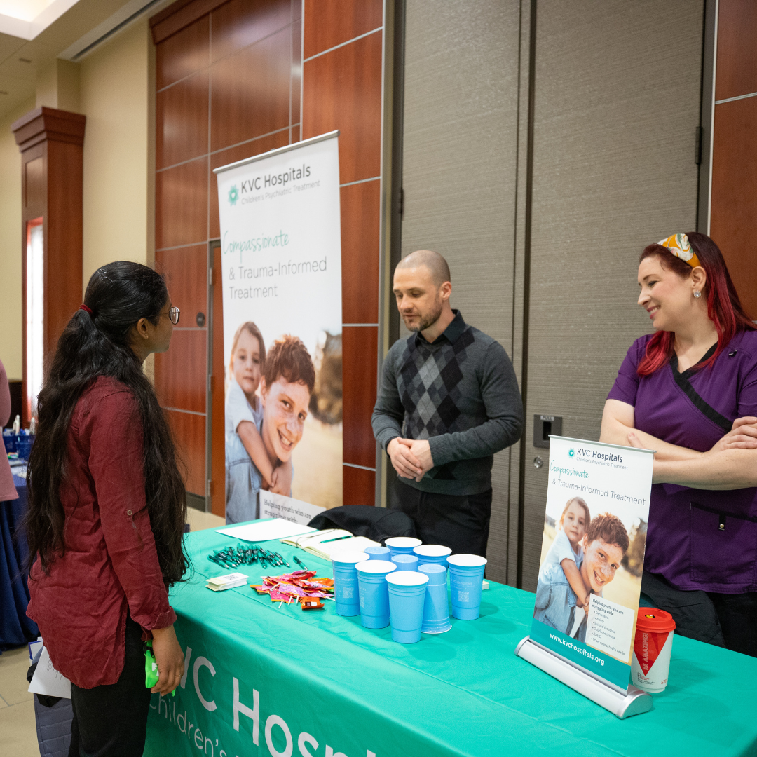 A student talks with two representatives at a career fair booth, with a company banner and table display in the background.