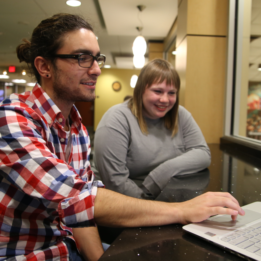 Two students at a laptop in a campus lounge, one pointing at the screen while the other looks on and smiles, with a pool table and seating area visible in the background.