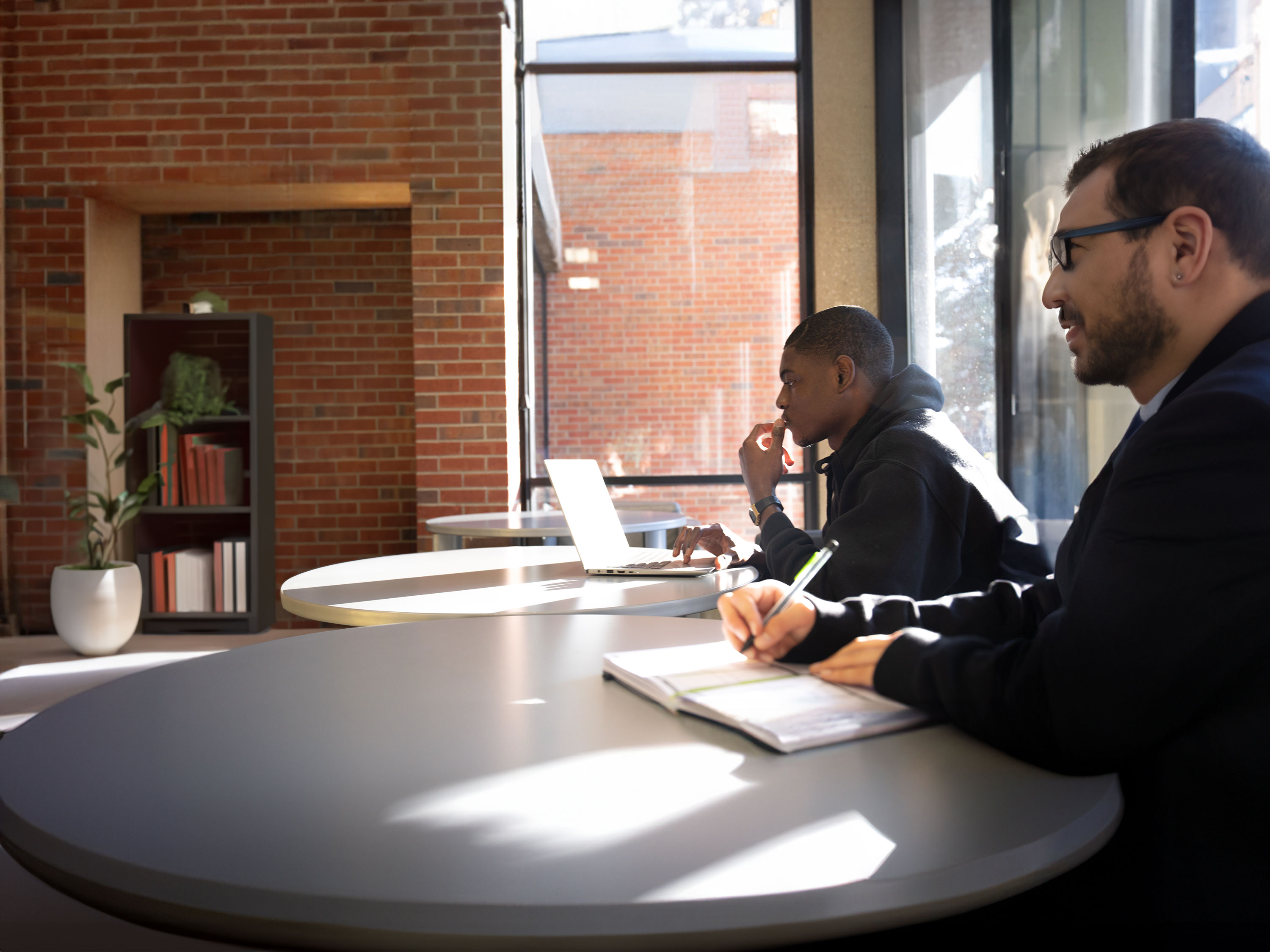 A student working on their laptop and sitting next to another person writing in a notebook.