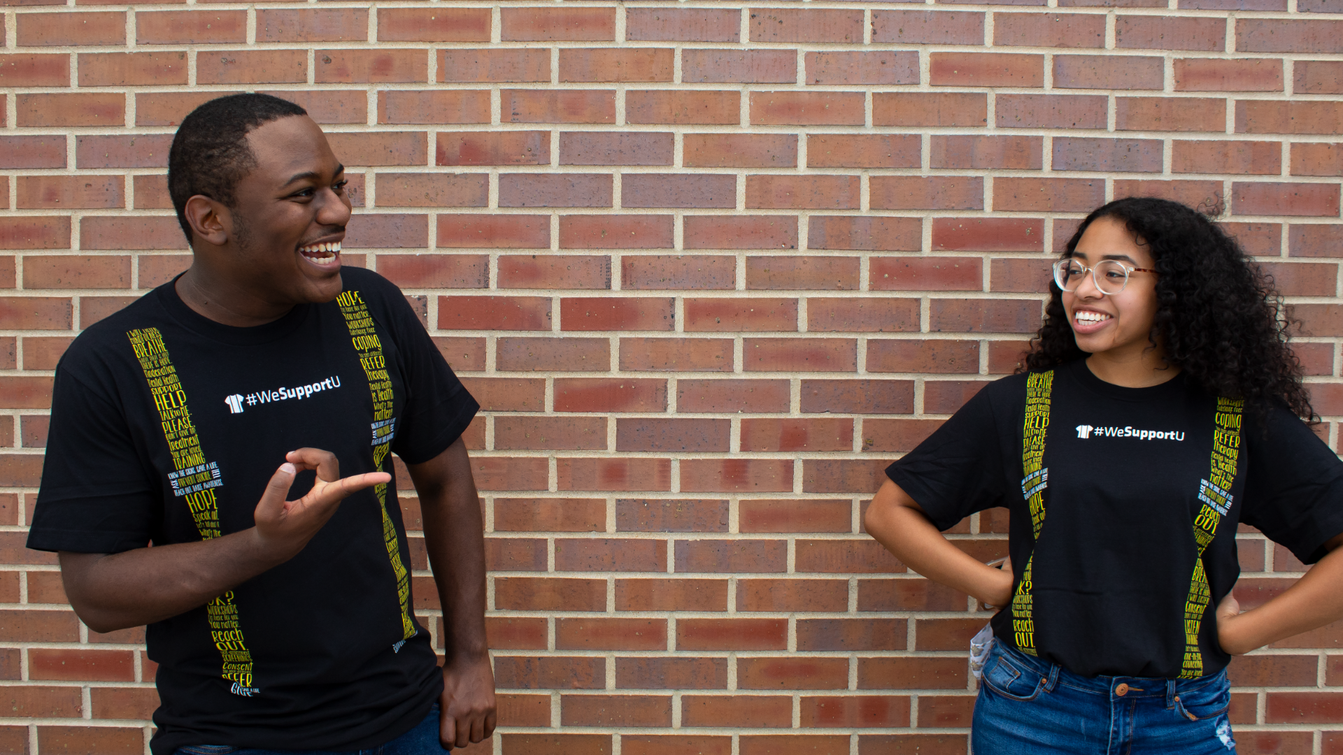 Two individuals, one who is tall with close buzzed hair and the other who is short with long curly hair laughing. Both are wearing Suspenders4Hope shirts.