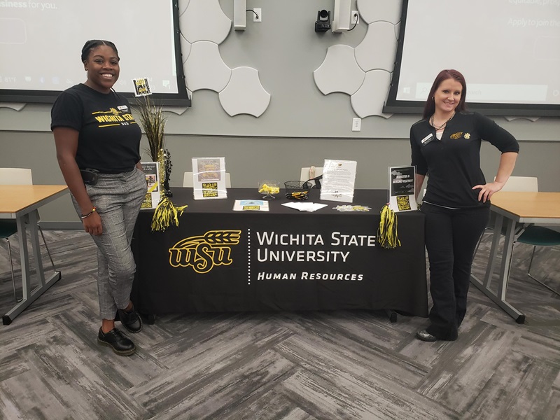 Two representatives of the Human Resources team standing at an information table during an event. 