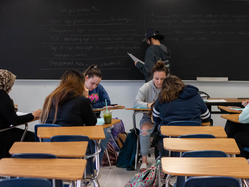 Four students in a classroom with their desks turned to face each other. Another student writes at the board behind them.