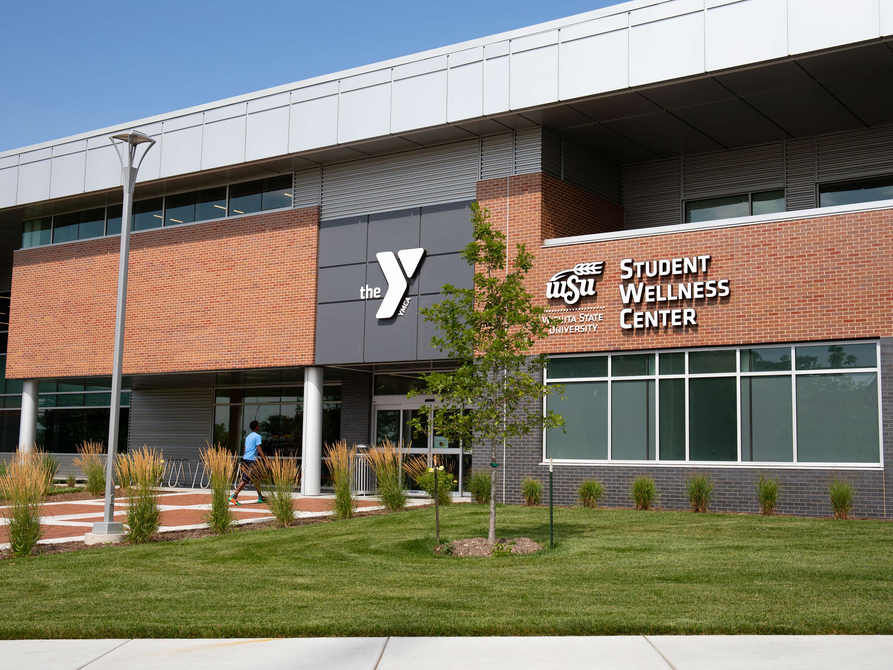 A student walks into the WSU Student Wellness Center. Green landscaping is visible in the foreground and the building's brick facade is visible in the background.