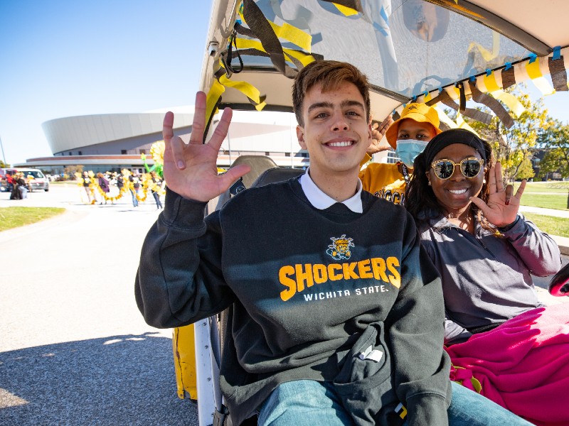 Homecoming parade participants ride in a golf car near Koch Arena