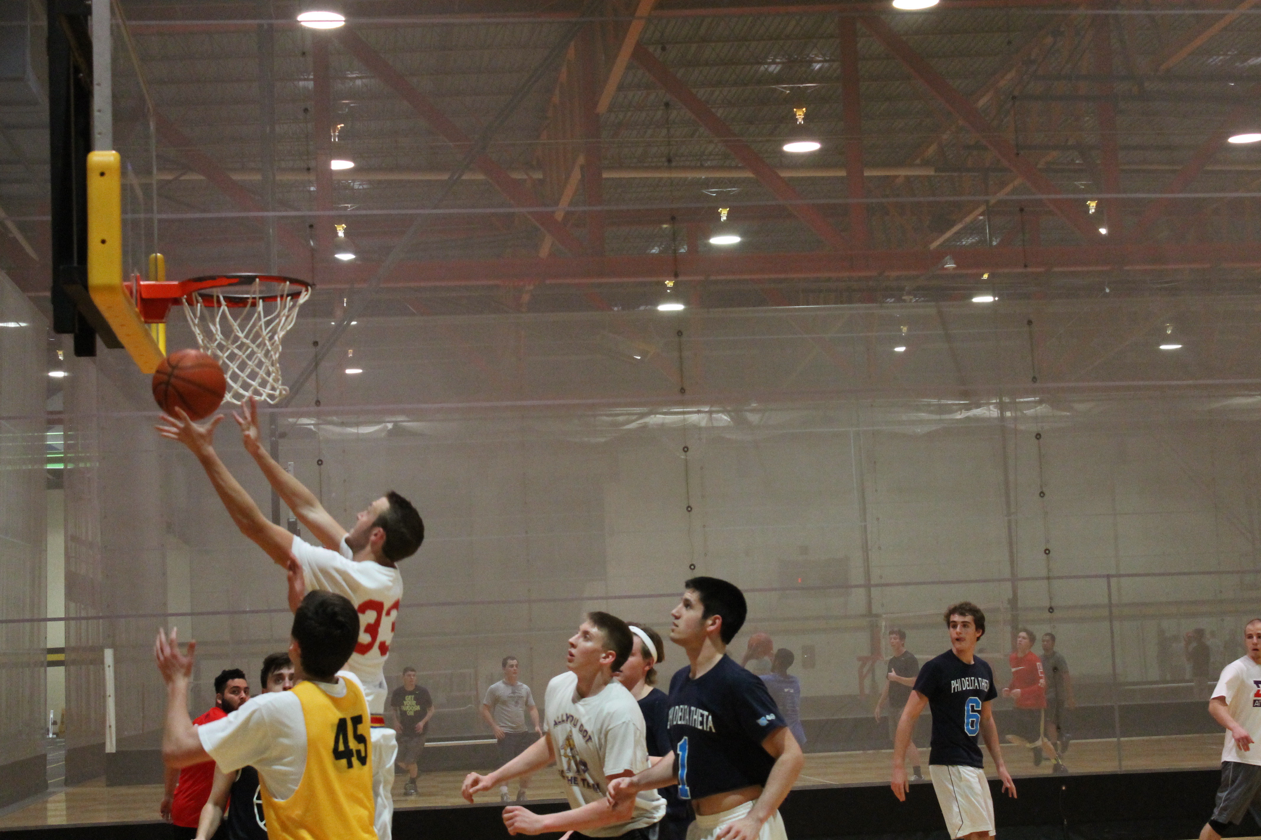 Basketball game in upstairs Heskett Center gym. One man is jumping up to make a basket. The backboard is perpendicular to the camera. other players are gathered near the basket, watching the ball.