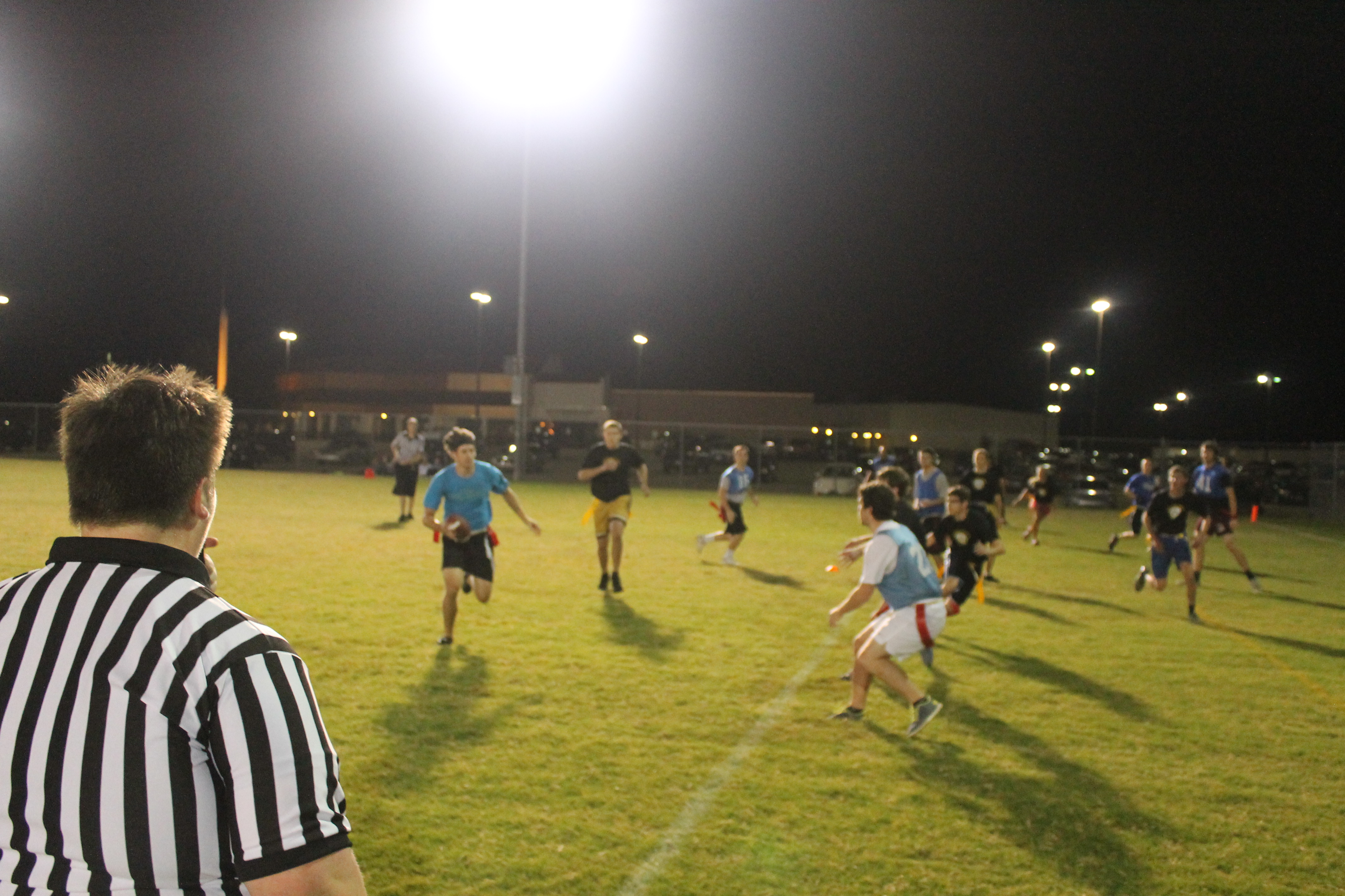 people playing flag football. camera is focused on the ref in the bottom left corner, who is facing away from us and toward the players.