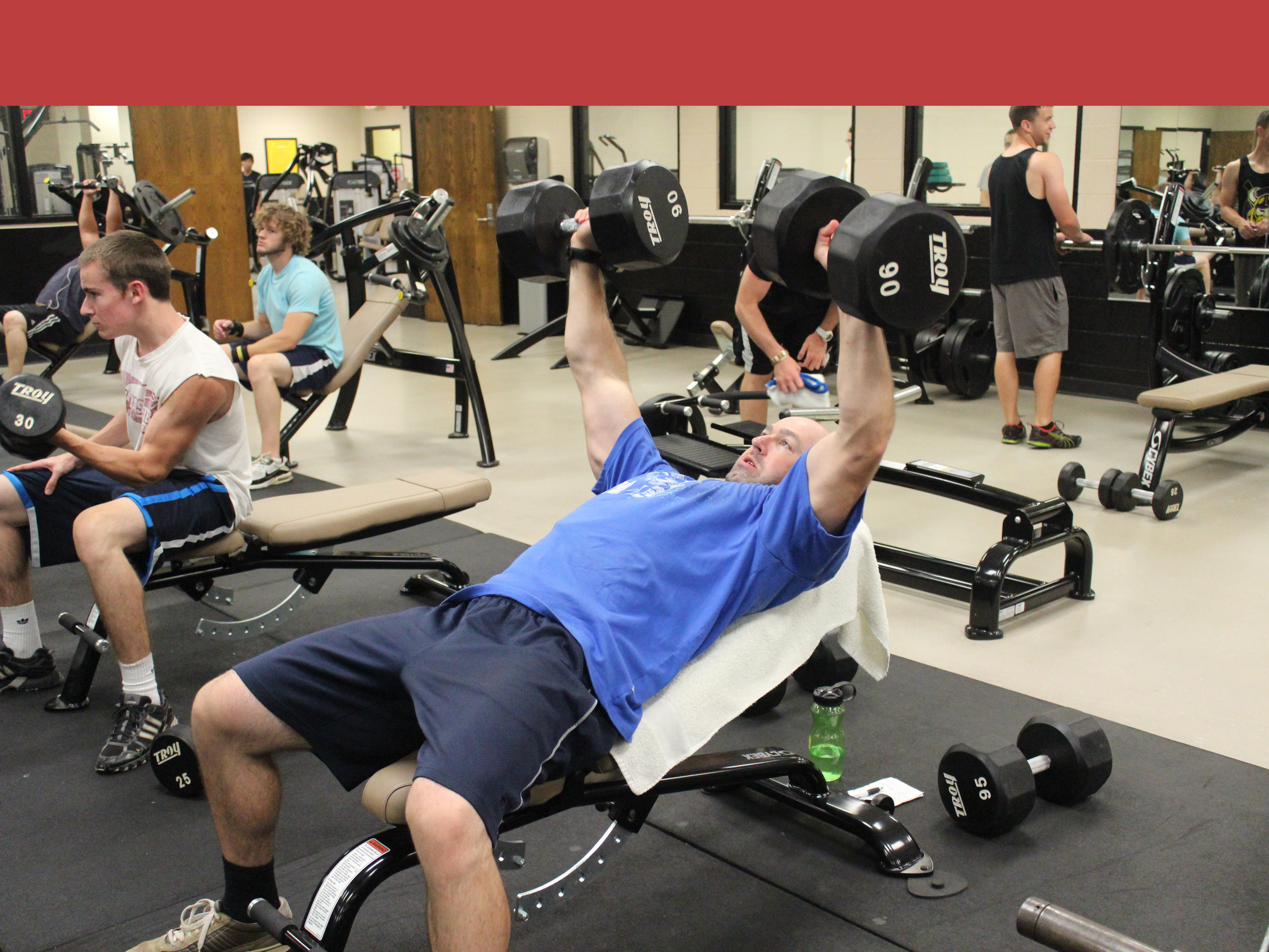 man doing dumbbell bench press in the weight room. lifting two 90's. several other weightlifters in the background.