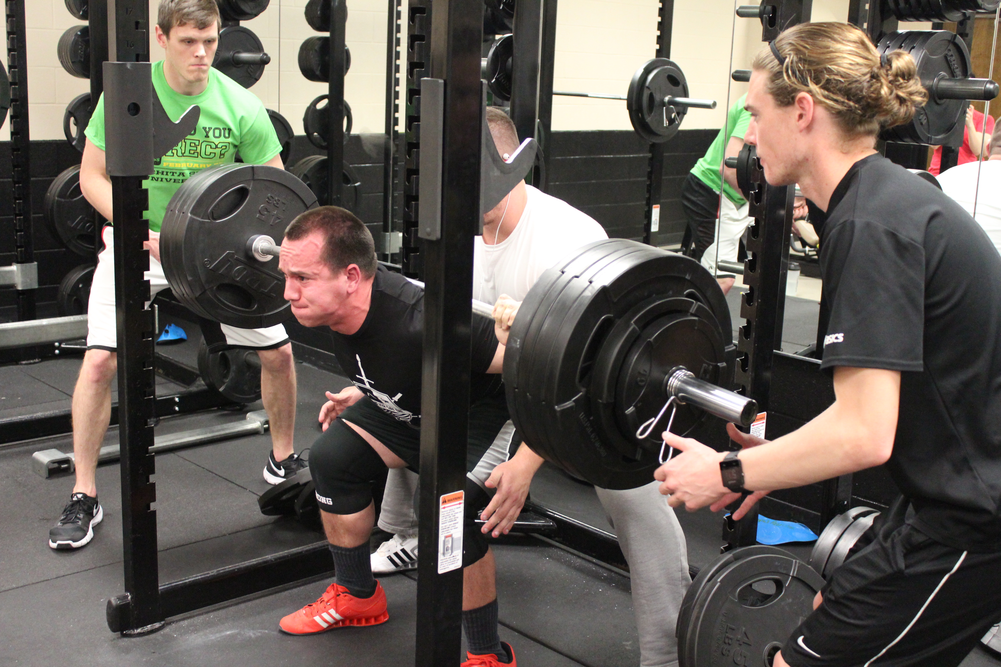 a man, red faced, straining under a bar stacked with 4 45 pound weights on each side. two other men are spotting him, one on each end of the bar