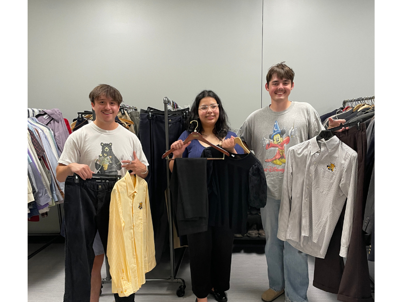 hree people stand in a clothing room, smiling at the camera while holding coordinated outfits on hangers. The person on the left holds a pair of dark pants and a light yellow button-down shirt with a small embroidered bee logo. The person in the center holds an all-black outfit with black pants and a black top. The person on the right holds a gray-and-white striped button-down shirt with the same bee logo. Clothing racks filled with shirts and pants surround them in the background.