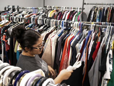 A person browses a densely packed clothing rack, looking through garments on hangers in what appears to be a campus clothing closet or thrift space. Rows of shirts, jackets, dresses, and pants in a variety of colors fill multiple racks, emphasizing the wide selection of available clothing.