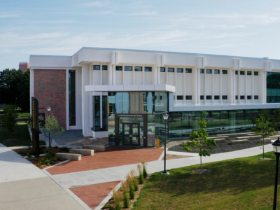 A modern two-story campus building with a white exterior, large glass windows, and a glass-front entrance sits along landscaped walkways and green lawns. Young trees, lampposts, and brick pathways surround the building under a clear blue sky. A vertical sign near the entrance reads "Student Success Center," and the entrance canopy is labeled "Lair Foundation Entrance."