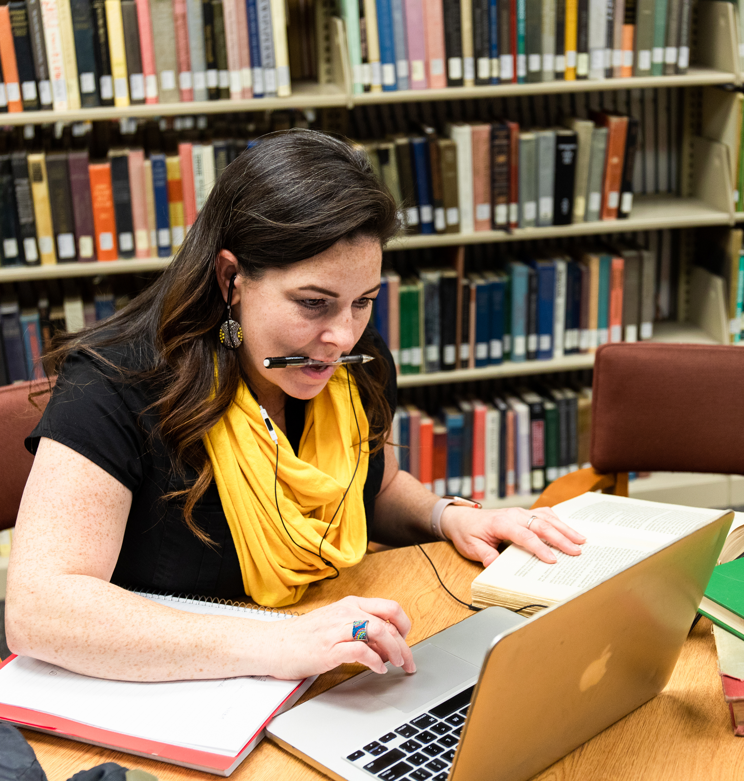 Adult learner holding a mechanical pencil in her mouth with her hand on the trackpad of her laptop in the library.
