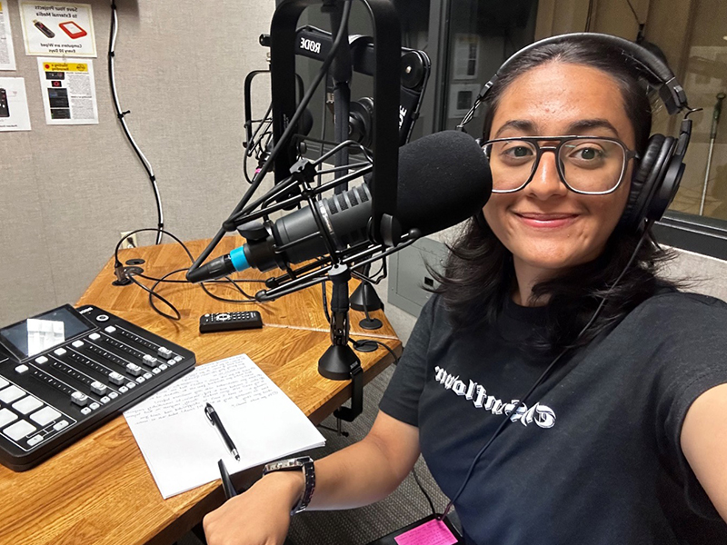 Image of Nidhi Shenoy behind a mic in a recording book with a notebook open in front of her. She's wearing a Sunflower t-shirt.
