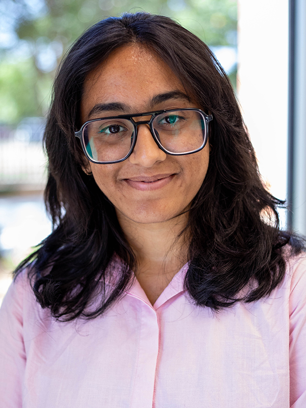 Professional photo of Nidhi Shenoy. She's wearing glasses and a pink button shirt.