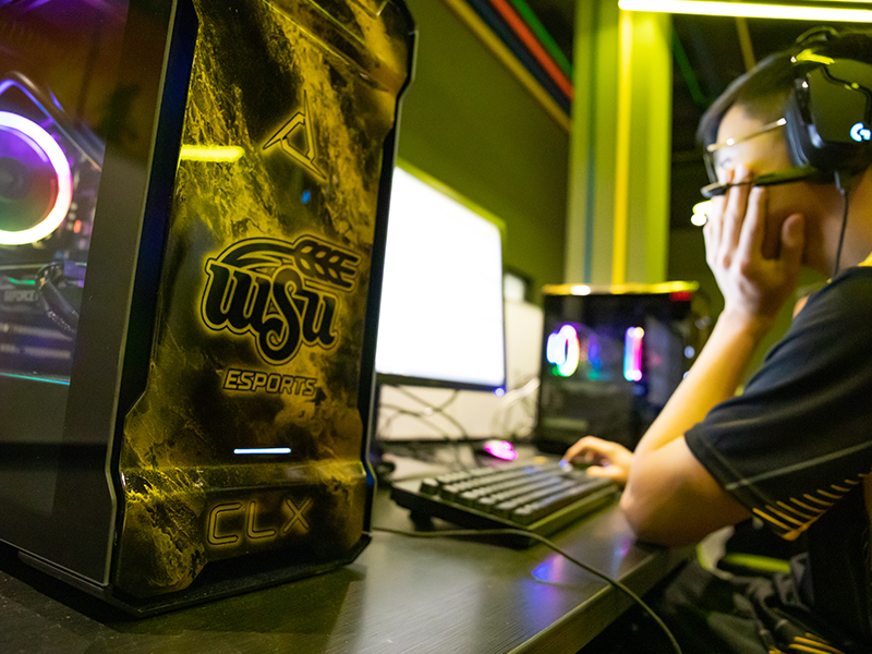 A closeup of a gaming consule that says WSU esports. In the background is a student playing a video game. 