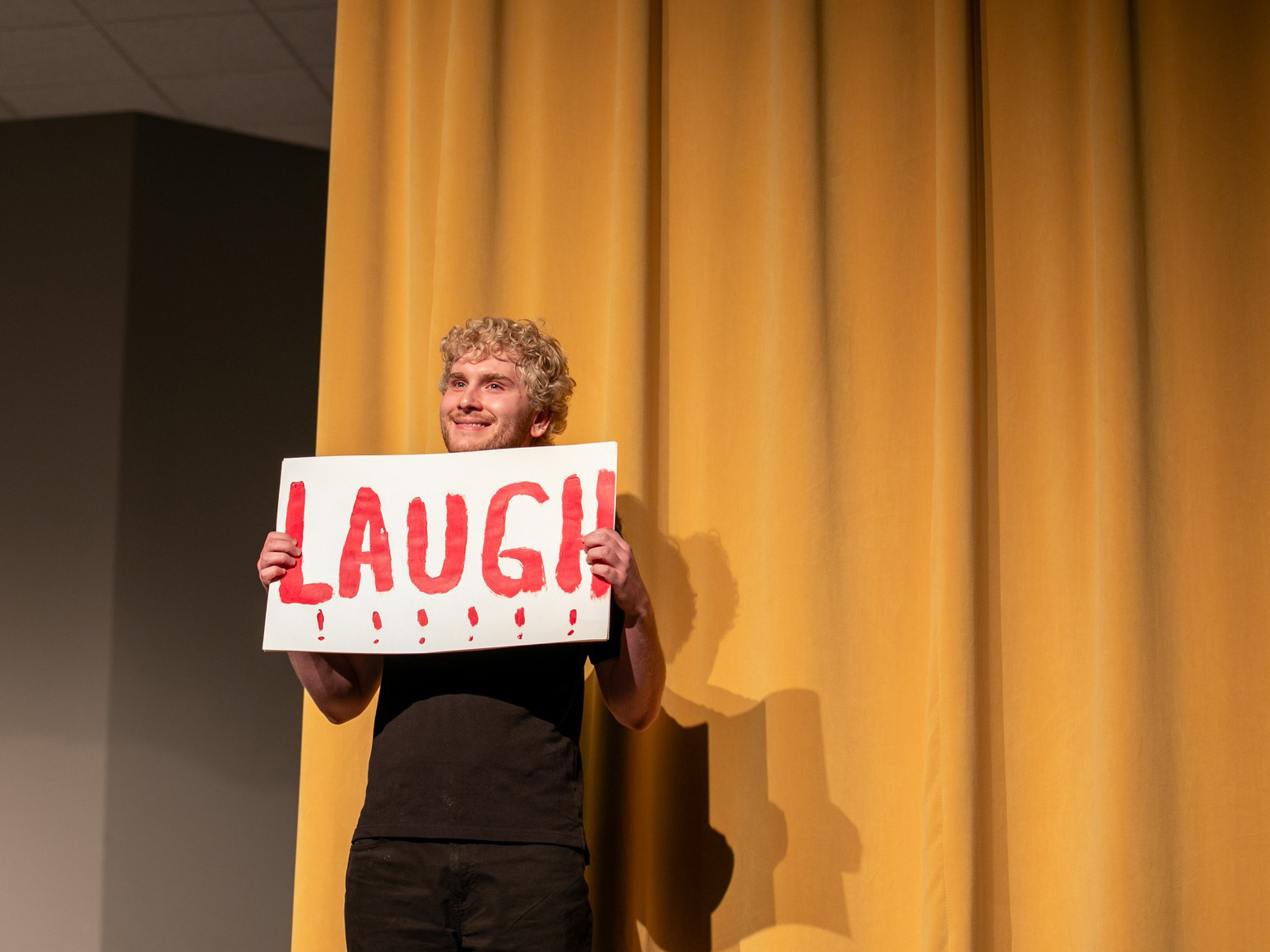 A student holding a sign that reads, "Laugh," poses in front of a yellow curtain.