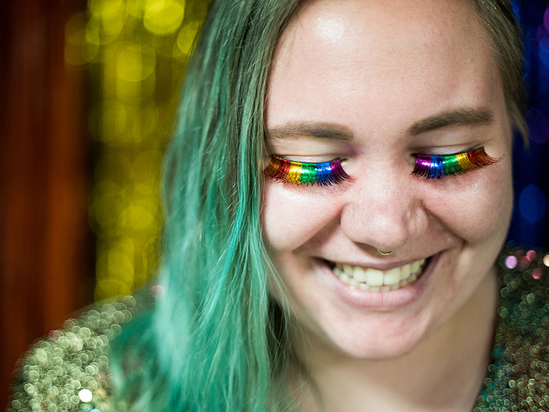 Student with rainbow colored eye lashes and bright hair posing for the camera at an LGBTQ+ event.